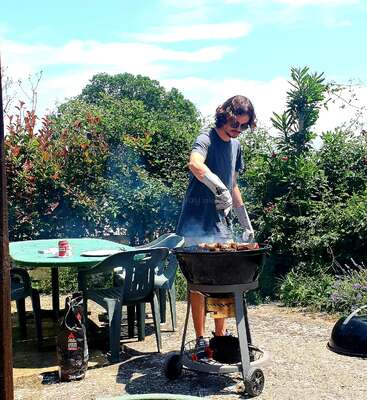 A man barbecues outdoors on a sunny day, wearing gloves and sunglasses. Smoke rises from the grill. Green table, chairs, and lush garden surround him.
