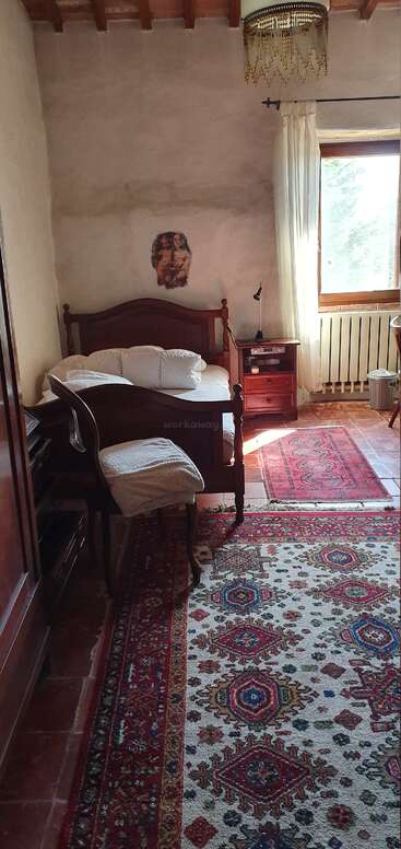 Cozy bedroom with wooden furniture, patterned rugs, sunlight filtering through a window, cream walls, art above the bed, chandelier, bedside table, and inviting atmosphere.