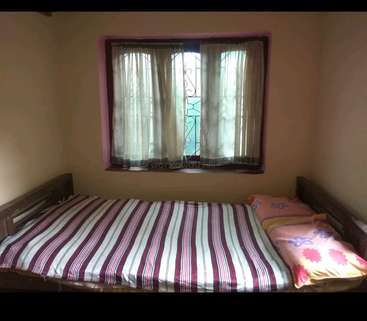 The image depicts a small bedroom with a single bed, featuring a striped duvet cover and two pillows, accompanied by a window with white curtains and a decorative metal grate.