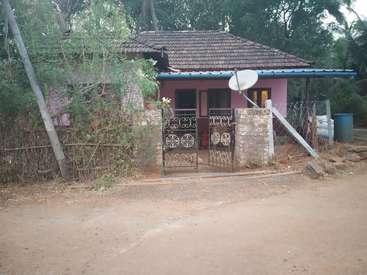 The image depicts a small, pink house with a tiled roof, surrounded by trees and a dirt road, featuring a satellite dish and a blue water tank on the right side.