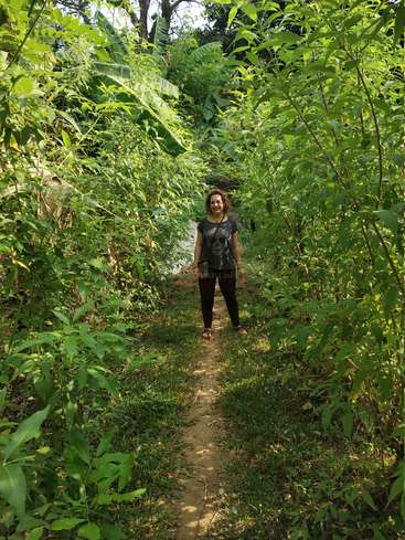 A woman stands on a dirt path in a lush, tropical forest, surrounded by vibrant greenery and trees, with sunlight filtering through the foliage.