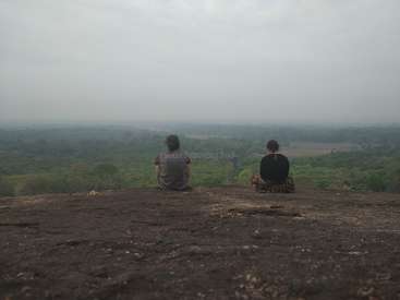 Two people sit on a rocky outcrop, gazing out at a lush green landscape under a gray sky, with trees and fields visible in the distance.