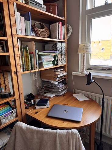 Un espace de travail chaleureux, avec des étagères en bois remplies de livres, de paniers et d'articles de papeterie. Un ordinateur portable est posé sur un bureau bien rangé, près d'une fenêtre laissant entrer la lumière naturelle.