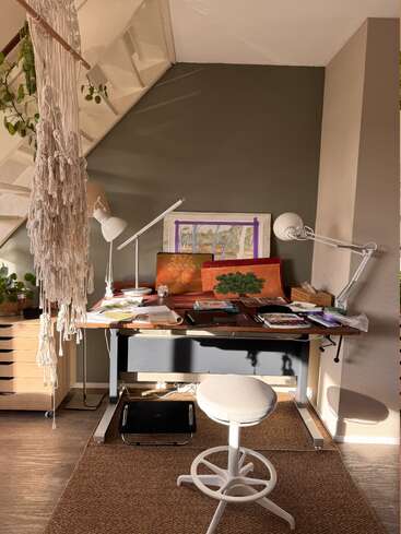 Un coin de travail chaleureux sous l'escalier, avec un bureau en bois, du matériel artistique, des carnets de croquis, deux lampes, des plantes, un tabouret blanc, un tapis tissé et une décoration murale en macramé.