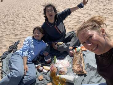Drei Freunde genießen ein sonniges Picknick am Strand; sie sitzen lächelnd und entspannt auf einer Decke, während Essen, Getränke und Sandalen im Sand verstreut liegen.