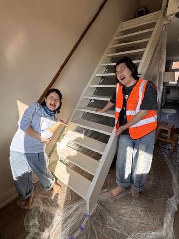 Zwei lächelnde Menschen präsentieren stolz eine frisch gestrichene Treppe. Einer trägt eine orangefarbene Warnweste. Der Boden und die Treppe sind zum Schutz während der Renovierungsarbeiten mit Plastikfolie abgedeckt.