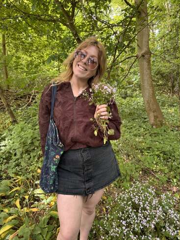 Eine lächelnde Frau steht in einem grünen Wald, trägt eine Sonnenbrille, eine weinrote Jacke und einen Jeansrock und hält einen Strauß Wildblumen und eine Tasche mit Blumenmuster in der Hand.