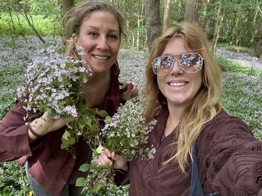 Zwei Frauen lächeln in einem blumenreichen Wald. Beide halten Sträuße aus violetten Wildblumen in den Händen und tragen weinrote Oberteile. Eine trägt eine verspiegelte Sonnenbrille. Eine fröhliche, naturverbundene Szene.