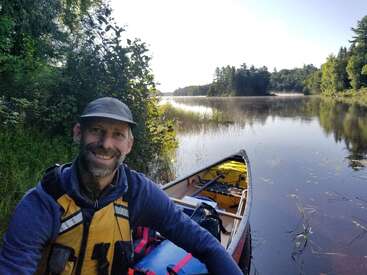 A man in outdoor gear smiles beside a loaded canoe on a calm, reflective river surrounded by lush green trees under a bright, clear sky.