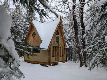 A cozy wooden cabin with a steep roof sits amidst a snowy forest, surrounded by tall trees and untouched snow, creating a peaceful winter retreat.