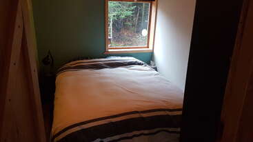 This cozy bedroom features a neatly made bed with striped bedding, a green accent wall, a reading lamp, and a window overlooking a peaceful forest scene.
