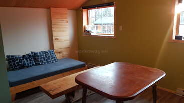 This cozy room features a wooden ceiling, olive green walls, a cushioned bench with patterned pillows, wooden coffee table, square dining table, and two windows.
