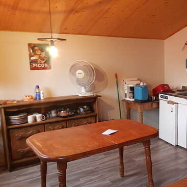 A cozy kitchen with wooden furniture, a fan, countertop appliances, a Picon poster on the wall, and a table with a note, under warm lighting.