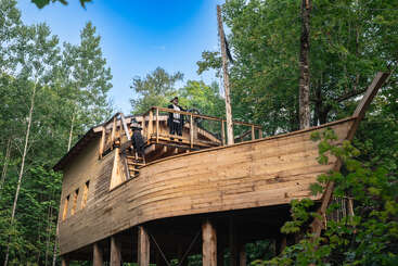 Ein hölzernes Baumhaus im Stil eines Piratenschiffs steht zwischen grünen Bäumen. Zwei Personen in Piratenkostümen stehen auf dem Deck und genießen die skurrile Umgebung.