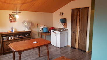 Une cuisine confortable avec des meubles en bois, un ventilateur, des appareils électroménagers de comptoir, un poster de Picon au mur et une table avec une note, sous un éclairage chaleureux.