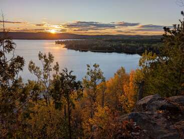 Un magnifique coucher de soleil baigne d'une lumière dorée un lac serein, entouré d'arbres aux couleurs chatoyantes de l'automne et de collines lointaines, sous un ciel partiellement nuageux. Une scène paisible.