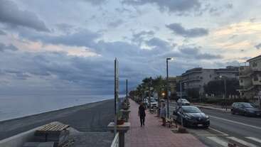 Ein ruhiger Küstenweg in der Abenddämmerung, gesäumt von Palmen, geparkten Autos und modernen Gebäuden. Der bewölkte Himmel spiegelt ein sanftes Licht über dem ruhigen Meer und der stillen Straße.