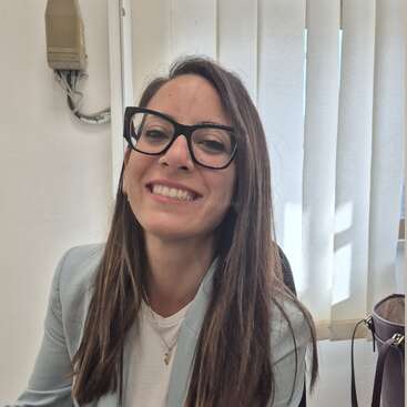 Uma mulher de cabelos castanhos compridos e óculos está sorrindo. Ela veste um blazer claro e uma camisa branca e está sentada dentro de casa, perto de uma janela com persianas verticais.