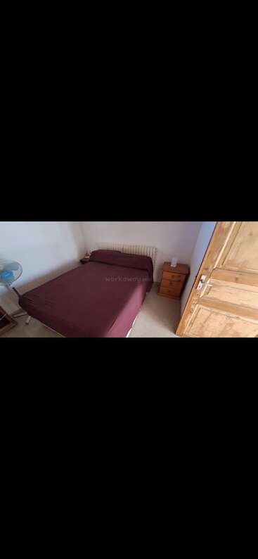 Dieses Bild zeigt ein einfaches Schlafzimmer mit einem kastanienbraunen Bett, einem Kleiderschrank aus Holz, zwei Nachttischen aus Holz, einem kleinen Ventilator und weißen Wänden. Minimalistische Einrichtung.