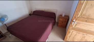 This image shows a simple bedroom with a maroon-covered bed, a wooden wardrobe, two wooden nightstands, a small fan, and white walls. Minimalist decor.