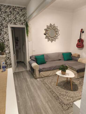 Uma sala de estar aconchegante com um papel de parede estampado como destaque, um sofá modular cinza, um espelho decorativo, uma guitarra pendurada na parede, uma mesinha de centro, uma planta de interior e um tapete de estilo vintage.