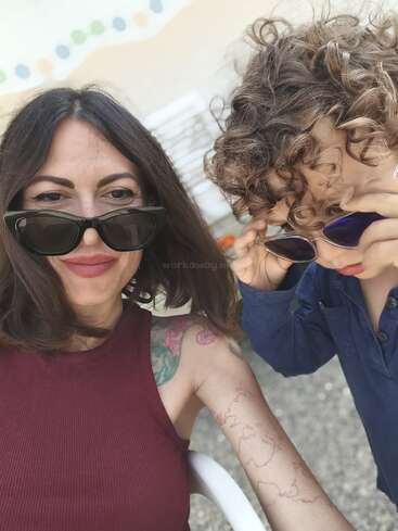 Uma mulher de cabelos castanhos longos e tatuagens está sentada ao lado de uma criança de cabelos cacheados. As duas estão usando óculos escuros, sorrindo e aproveitando juntas um momento ensolarado ao ar livre.