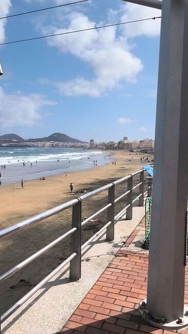 Uma cena ensolarada na praia com pessoas curtindo a areia e as ondas, montanhas ao fundo, prédios da cidade ao longo da costa e uma grade de metal em primeiro plano.