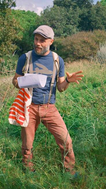 A man stands outdoors in tall grass, wearing a cap, suspenders, and carrying a striped bag. He gestures while holding papers, surrounded by greenery and sunlight.