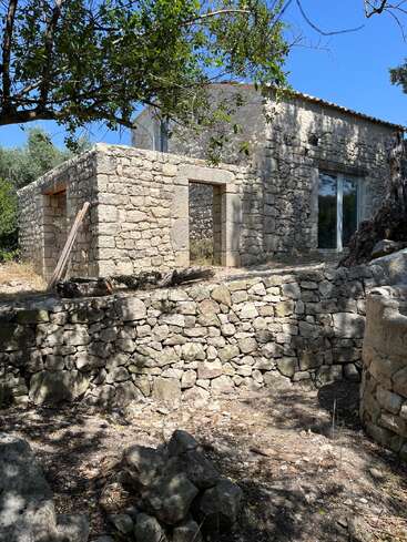 A rustic stone house stands under a blue sky, surrounded by trees and shaded areas. It features rough stone walls, arched doorways, and a tranquil atmosphere.