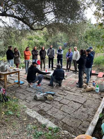 A group of people stands in a circle outdoors under a tree, apparently listening to two seated individuals. There's a table, tools, and a resting dog.