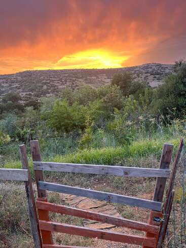 A wooden gate opens to a lush, green landscape with rolling hills. Above, a dramatic orange sunset illuminates the sky, creating a peaceful rural scene.