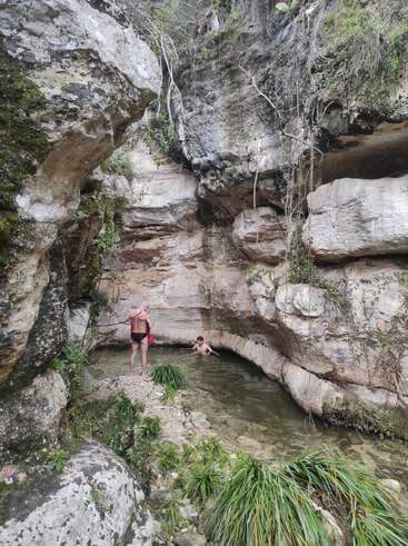 Two people are enjoying a small, natural pool surrounded by rugged rock formations, greenery, and hanging vines in a secluded, tranquil canyon environment.