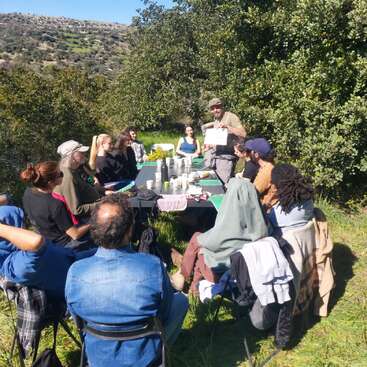 The image depicts a group of people gathered around a table in a serene outdoor setting, surrounded by trees and hills under a clear blue sky.