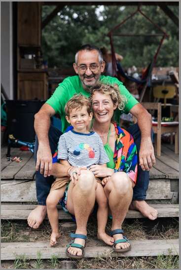 Una feliz familia de tres miembros está sentada muy junta en unos escalones de madera en el exterior. Sonríen cálidamente, disfrutando de un momento de relax, vestidos con ropa veraniega de colores vivos.