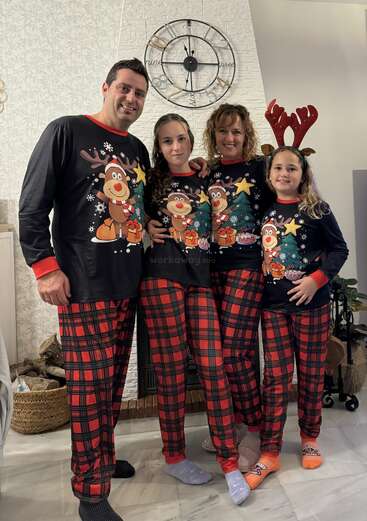 Una familia de cuatro miembros posa alegremente con pijamas navideños a juego con motivos de renos. Están en el interior, junto a un reloj, celebrando juntos unas acogedoras fiestas.