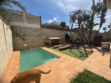Un acogedor jardín trasero cuenta con una pequeña piscina, un patio con baldosas, muebles de exterior, un columpio, altas palmeras, césped artificial, un muro de piedra y un perro. Un día soleado.