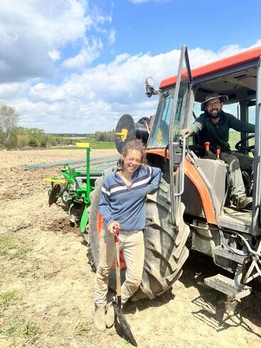 Dos personas en un día soleado: una está de pie, sonriendo, con una pala junto a un tractor, y la otra está sentada dentro, ambas vestidas para trabajar en el campo.