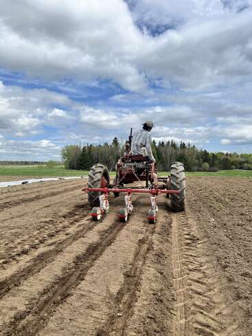 A farmer drives a tractor across a freshly plowed field, preparing soil for planting under a partly cloudy sky, surrounded by greenery and distant trees.