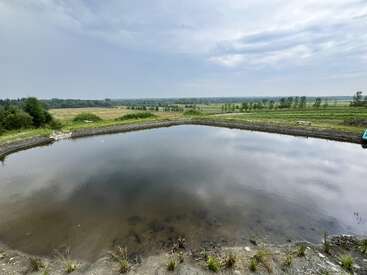 A small, calm pond with slightly muddy water sits in the middle of a grassy field, surrounded by open countryside under a mostly cloudy, bright sky.