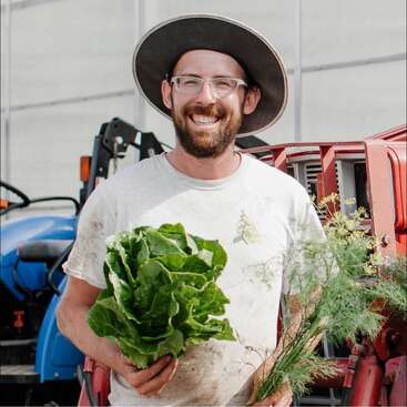 A smiling man wearing a wide-brimmed hat and glasses holds fresh lettuce and herbs, standing in front of a tractor, symbolizing farming and harvest joy.