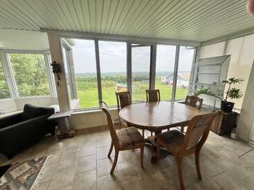 The image shows a sunroom with large windows, a wooden dining table with five chairs, a black armchair, tiled floor, green outdoor view, and natural lighting.
