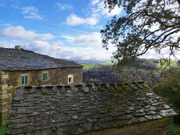 The image depicts a rustic stone house with a sloping roof, situated in a picturesque rural landscape with rolling hills and trees under a blue sky with white clouds.