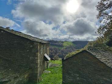 The image depicts a serene rural scene with stone buildings, a horse grazing in a lush green field, and a picturesque valley under a cloudy sky.