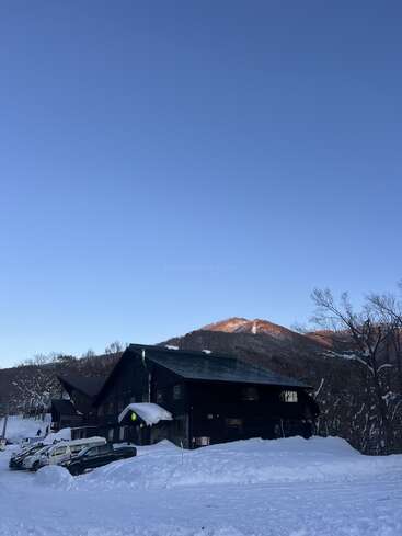 Eine Hütte aus dunklem Holz steht inmitten von Schnee, davor stehen mehrere Autos. Kahle Bäume und ein Berg, der im Licht der aufgehenden Sonne badet, runden das friedliche Winterbild ab.