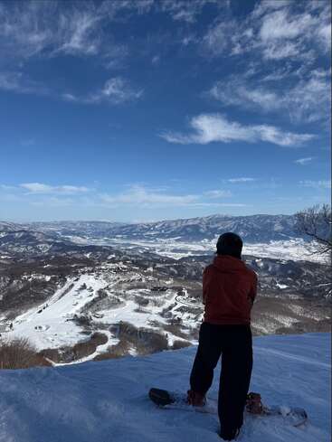 Ein Snowboarder steht am Rande eines schneebedeckten Berges und blickt auf ein weites, malerisches Tal mit fernen Gipfeln unter strahlend blauem Himmel und vereinzelten Wolken.