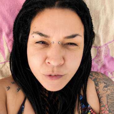 Une femme aux cheveux noirs tressés et au visage orné de piercings est allongée sur une serviette, partiellement tatouée, et regarde droit vers l'appareil photo, le front légèrement plissé.