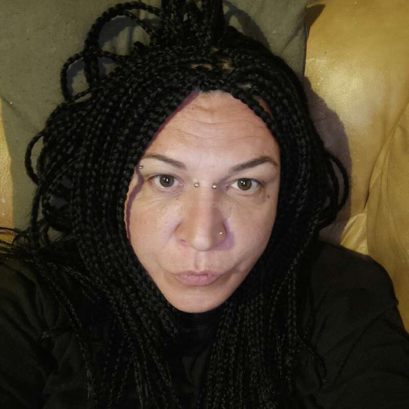 A woman with long black box braids and piercings above her eyebrows and nose looks directly at the camera, sitting on a couch with a neutral expression.