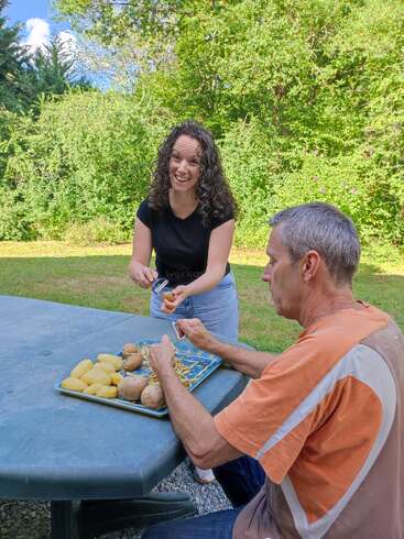 Una mujer y un hombre se sientan en una mesa al aire libre, pelando patatas juntos. Sonríen y disfrutan de su mutua compañía en un día soleado en plena naturaleza.