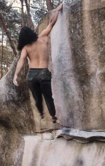 Una persona sin camiseta y con el pelo largo está practicando escalada en bloque al aire libre, tratando de alcanzar con una mano la cima de una gran pared rocosa rodeada de árboles.