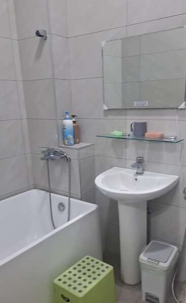 This image shows a compact bathroom with a bathtub, wall-mounted shower, pedestal sink, mirror, glass shelf, soap, mug, green stool, and a trash bin.